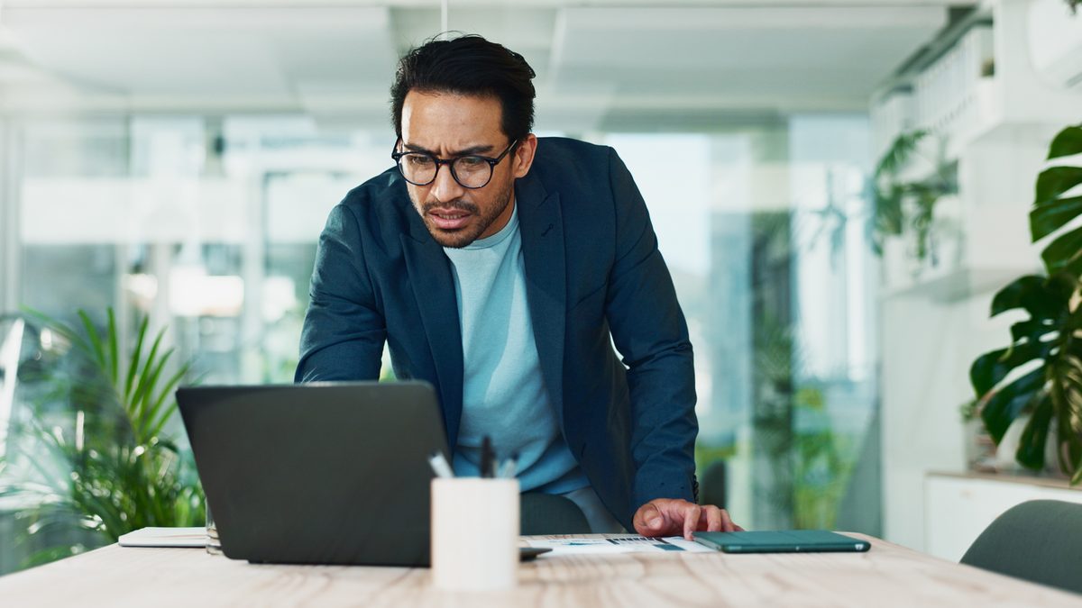 Small business owner in glasses leaning over a laptop at an office desk, reviewing small business homepage essentials on screen